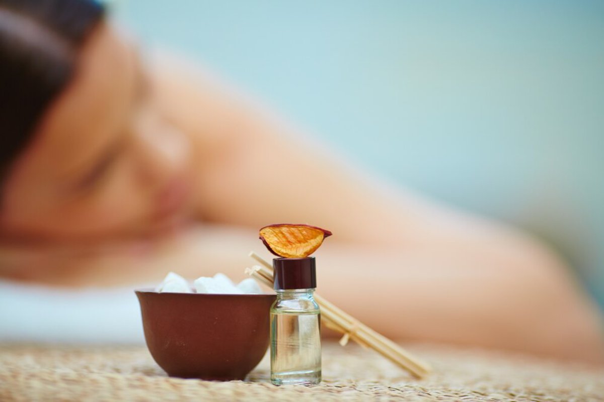 A small brown bowl of ice, a glass bottle of oil, and a dried orange slice lie beside a person receiving a massage in a serene setting.