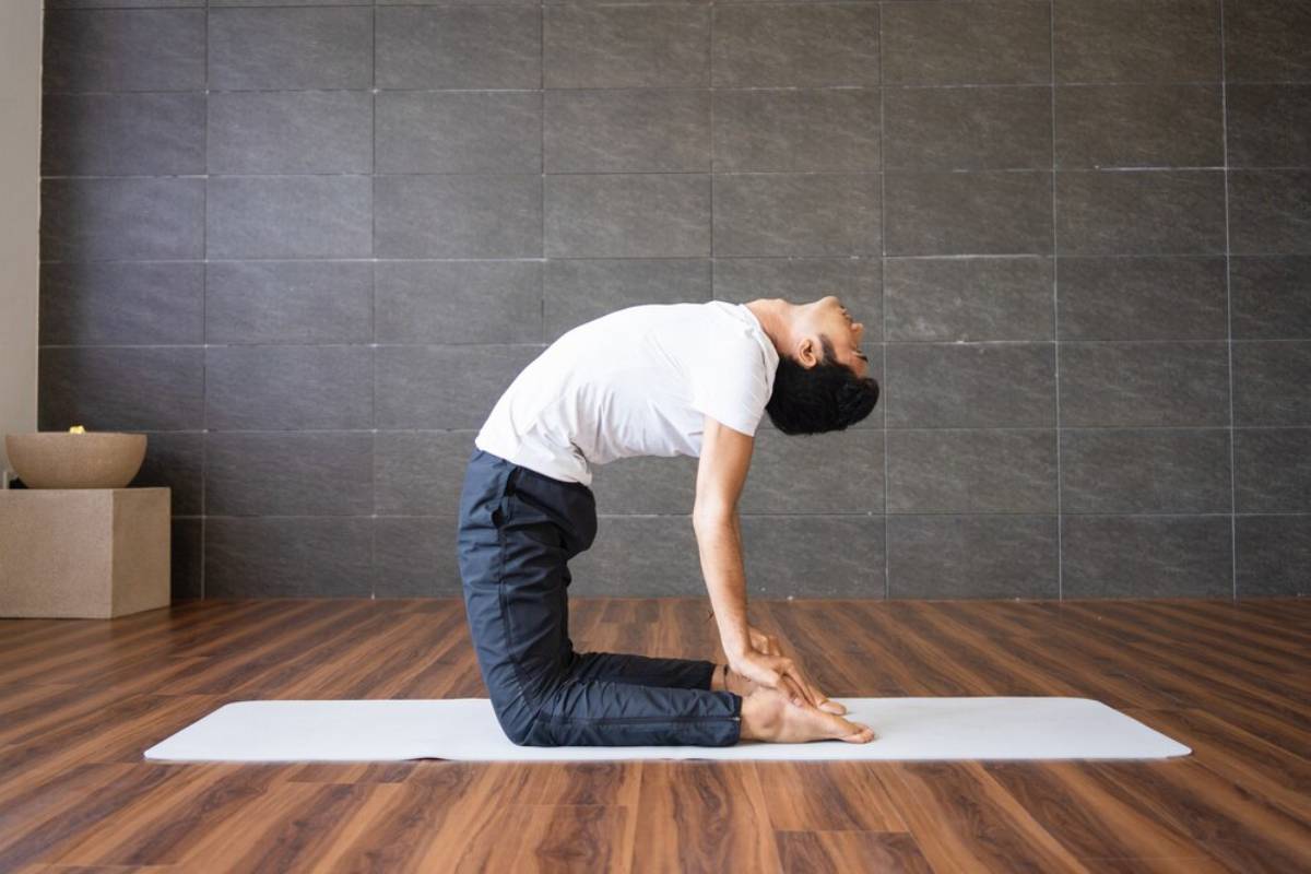 A person performs a yoga pose, Camel pose, with a backbend, on a mat, in a spacious, modern studio with stone walls.