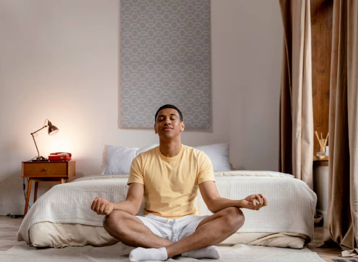 A person in a yellow t-shirt sits cross-legged on a bed, meditating in a serene, softly lit bedroom.