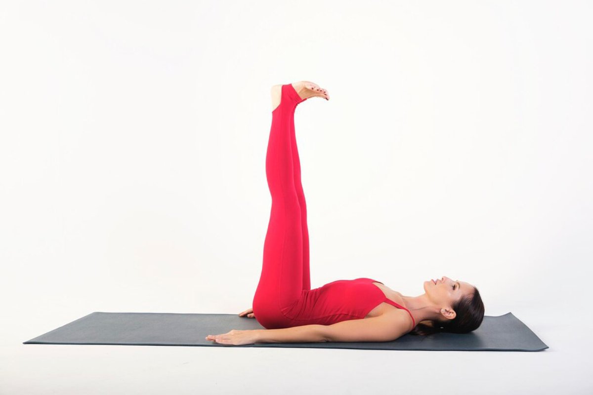 A person in a red outfit performs a yoga pose, lying on a mat with legs raised upwards against a white background.