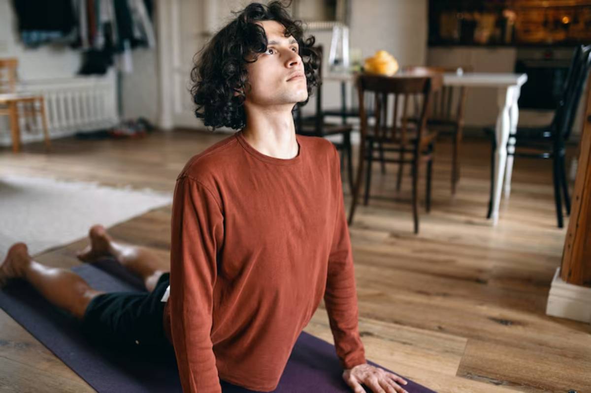 A person practices yoga in a cozy room with wooden floors and chairs in the background, focused on their pose.