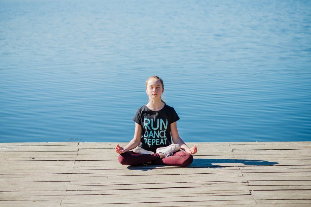 A person sits cross-legged on a wooden dock by a calm blue body of water, practicing meditation in a casual black shirt.
