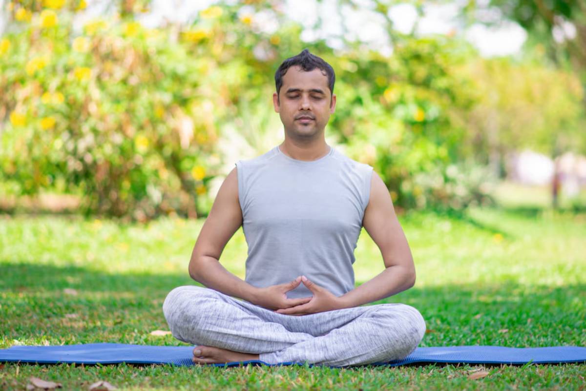 A person sits cross-legged on a yoga mat outdoors, practicing meditation amidst a lush green background.