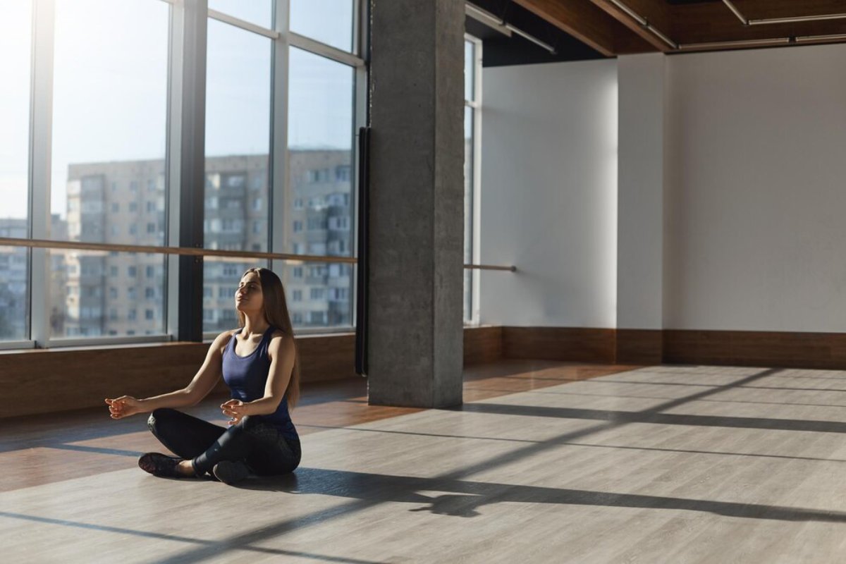 A person sits cross-legged in a sunlit studio, practicing meditation on a wooden floor with large windows showcasing an urban view.