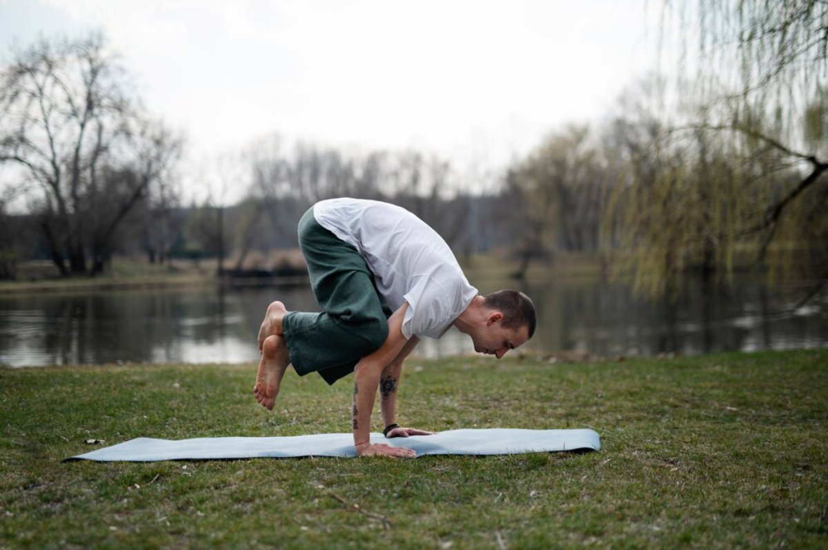 A person practicing yoga in an outdoor setting, balancing in a crow pose on a mat near a serene lake surrounded by trees.