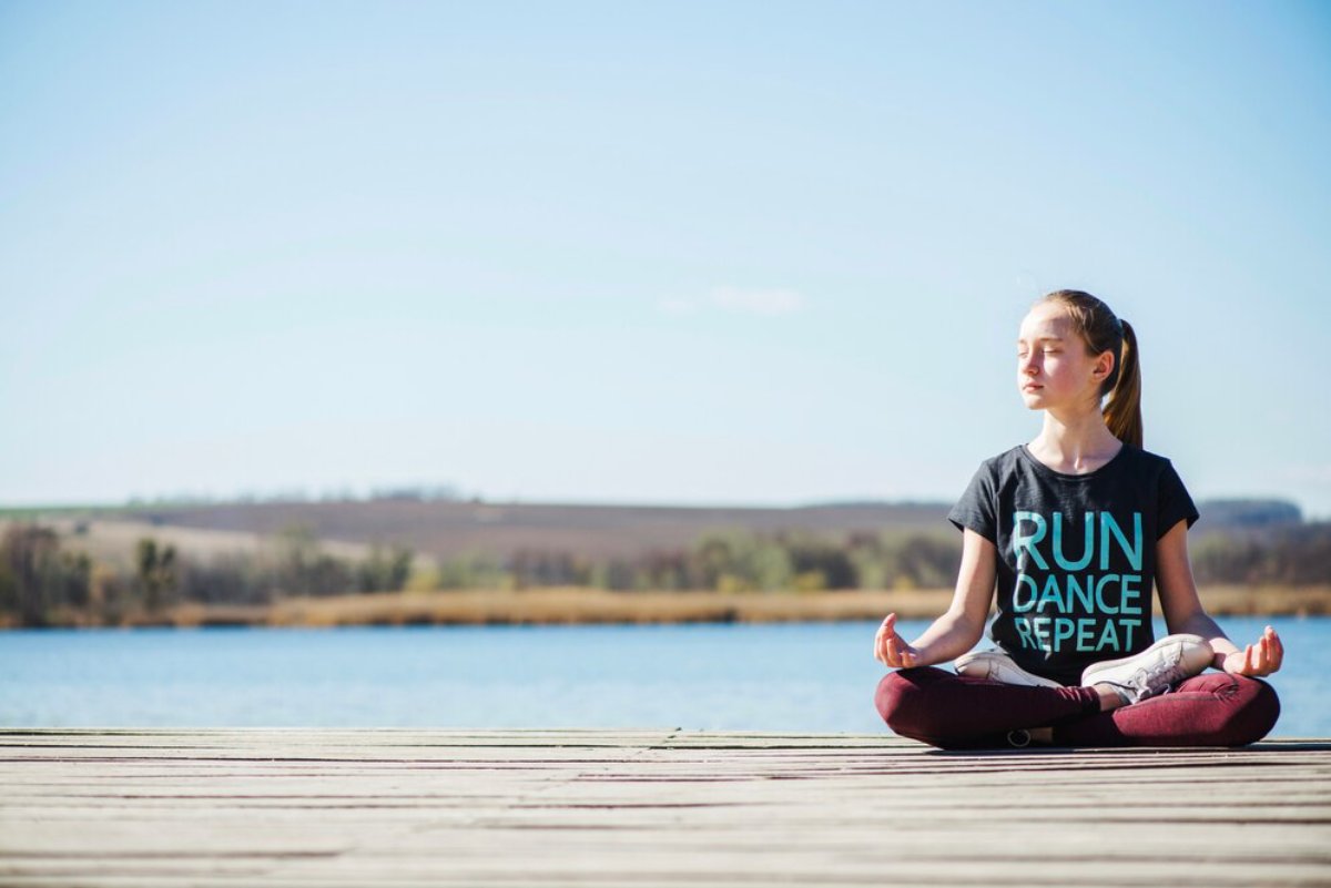 A girl sits cross-legged on a wooden dock by a calm lake, gazing into the distance on a clear, sunny day.