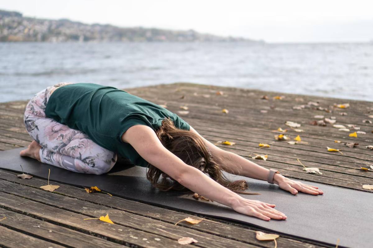 A woman practices yoga in Child's Pose on a mat by a lakeside, surrounded by autumn leaves and a tranquil water background.