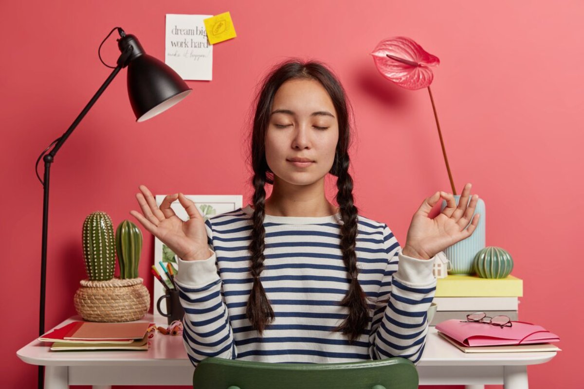 A person with braided hair sits at a colorful desk, practicing mindfulness amidst plants and playful decor against a pink backdrop.