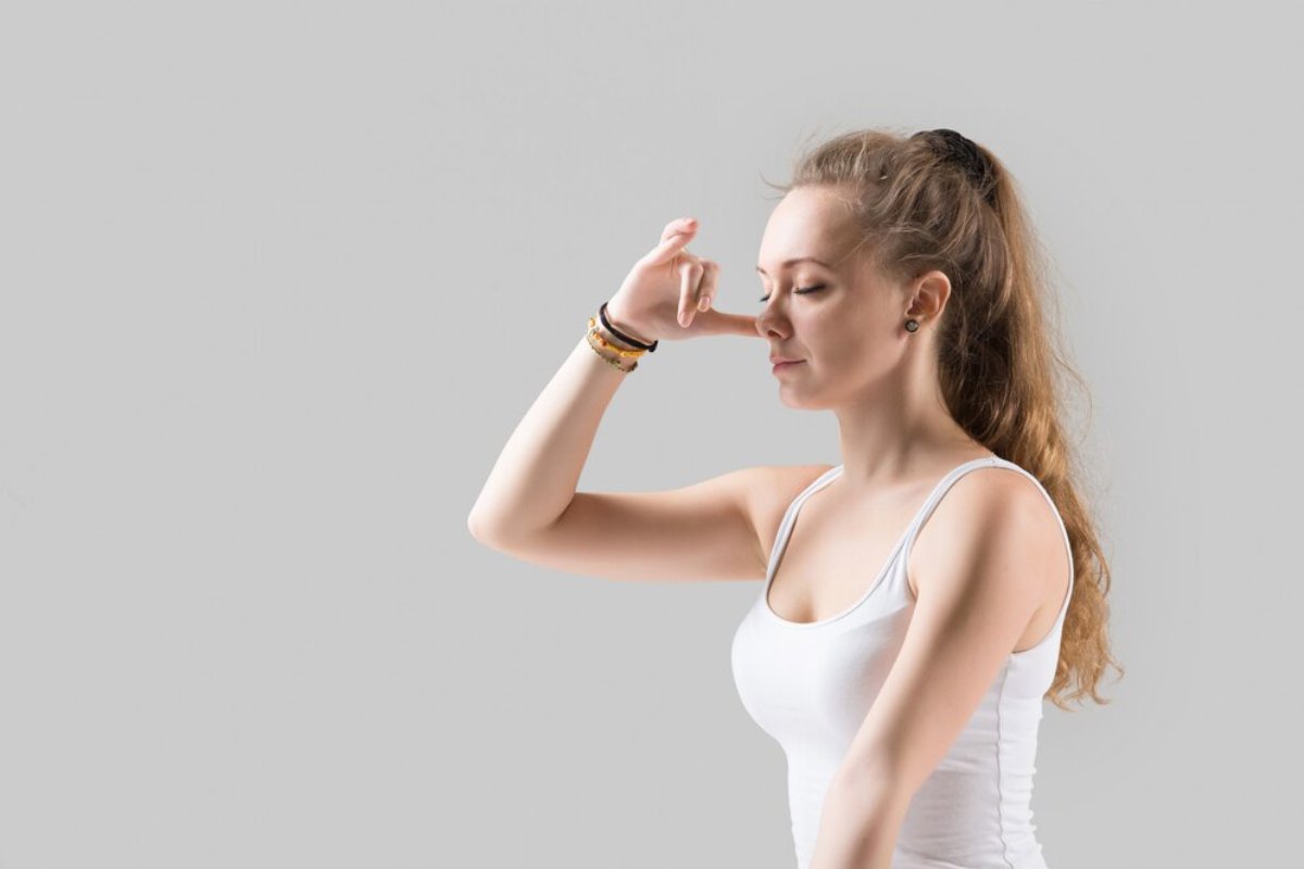 A young woman in a white tank top poses against a light gray background, confidently resting her hand beside her face.