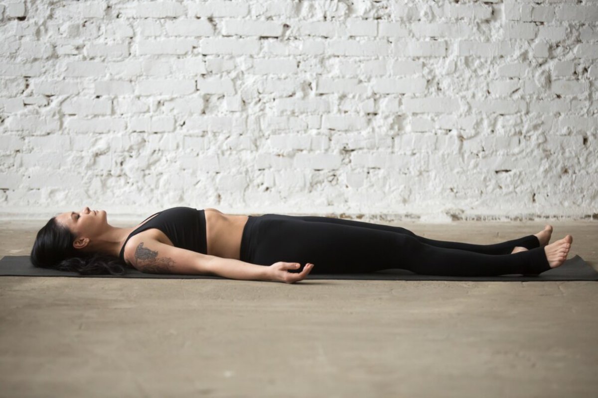 A person practices yoga in the shavasana pose on a mat, set against a textured white brick wall.