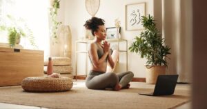 A person practicing yoga in a serene indoor space with plants, light streaming through windows, and a laptop nearby.