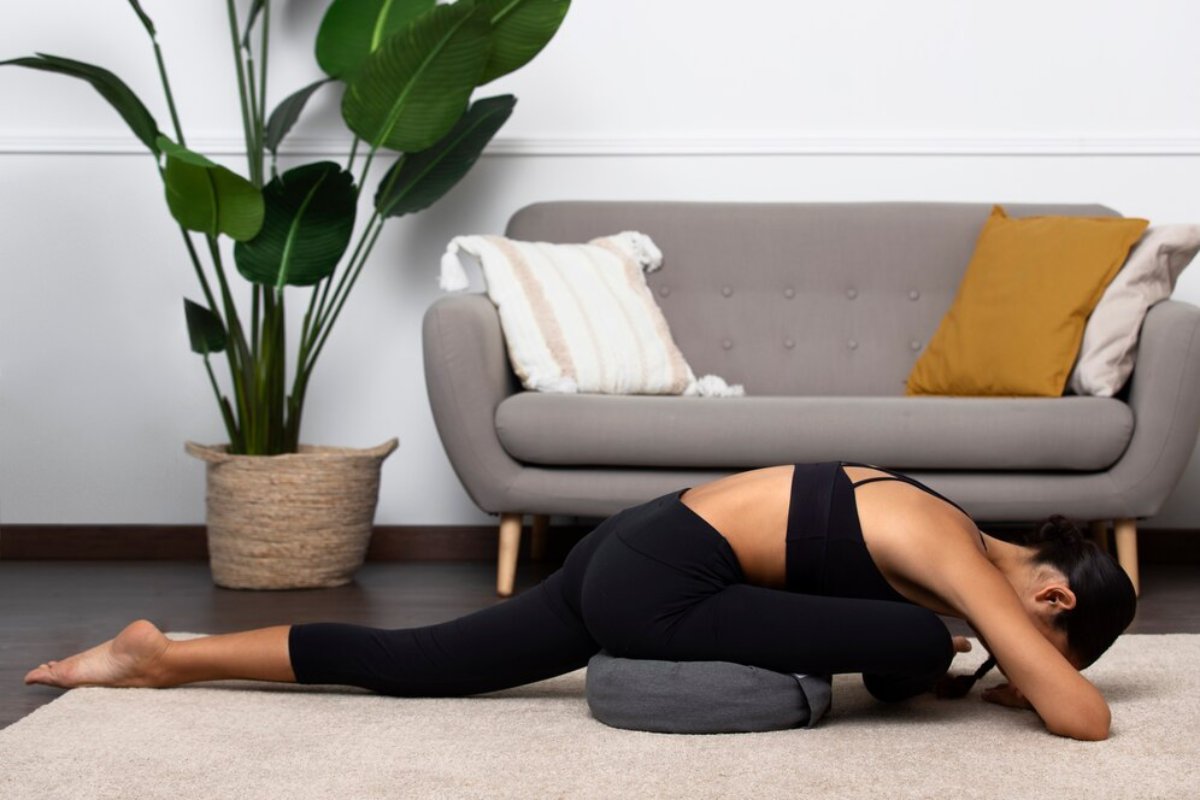 A woman in black activewear practices a yoga pose on a gray bolster, with a cozy living room and large plant in the background.