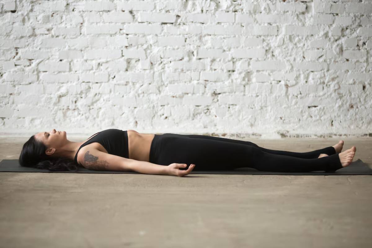 A person lies on a yoga mat in a relaxed position, wearing a black crop top and leggings, with a minimalist white brick wall in the background.
