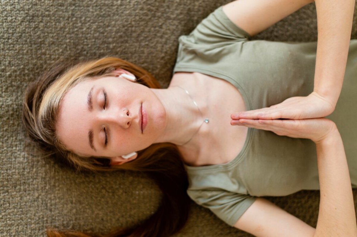 A woman lies on a soft surface, practicing mindfulness with hands together in a meditative pose, wearing a simple green shirt.