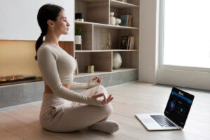 A person sits cross-legged on the floor in a modern room, meditating with a laptop playing soothing music nearby.