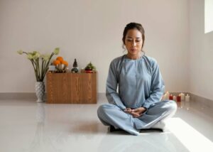 A person in a light blue outfit sits cross-legged in a serene room, surrounded by candles and a small altar with fruits and statues.