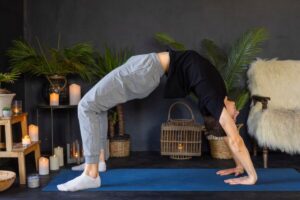 A person practicing yoga in a backbend position on a blue mat, surrounded by candles and plants in a serene indoor setting.