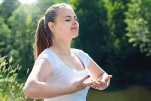 A woman in a white t-shirt gestures with her hands outdoors, surrounded by lush greenery and a serene background.