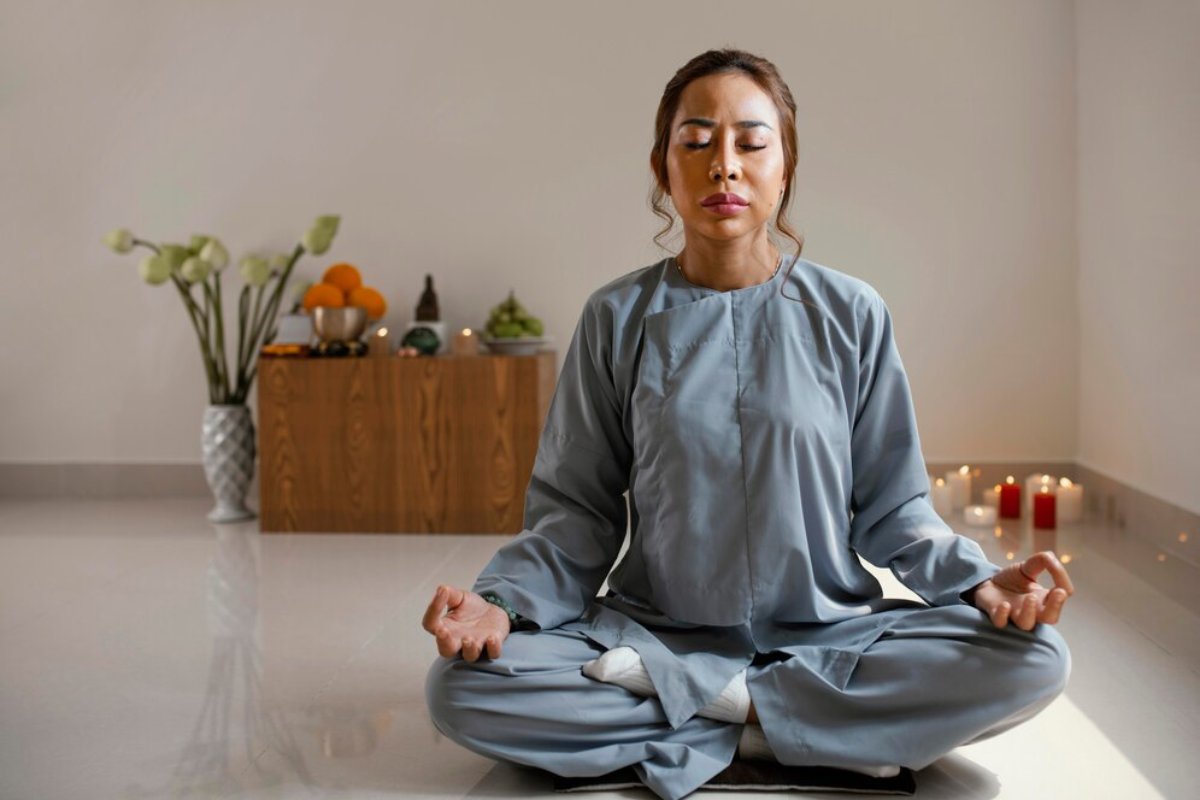 A person in a light blue robe meditates in a serene room with candles and decorative fruits on a wooden table in the background.