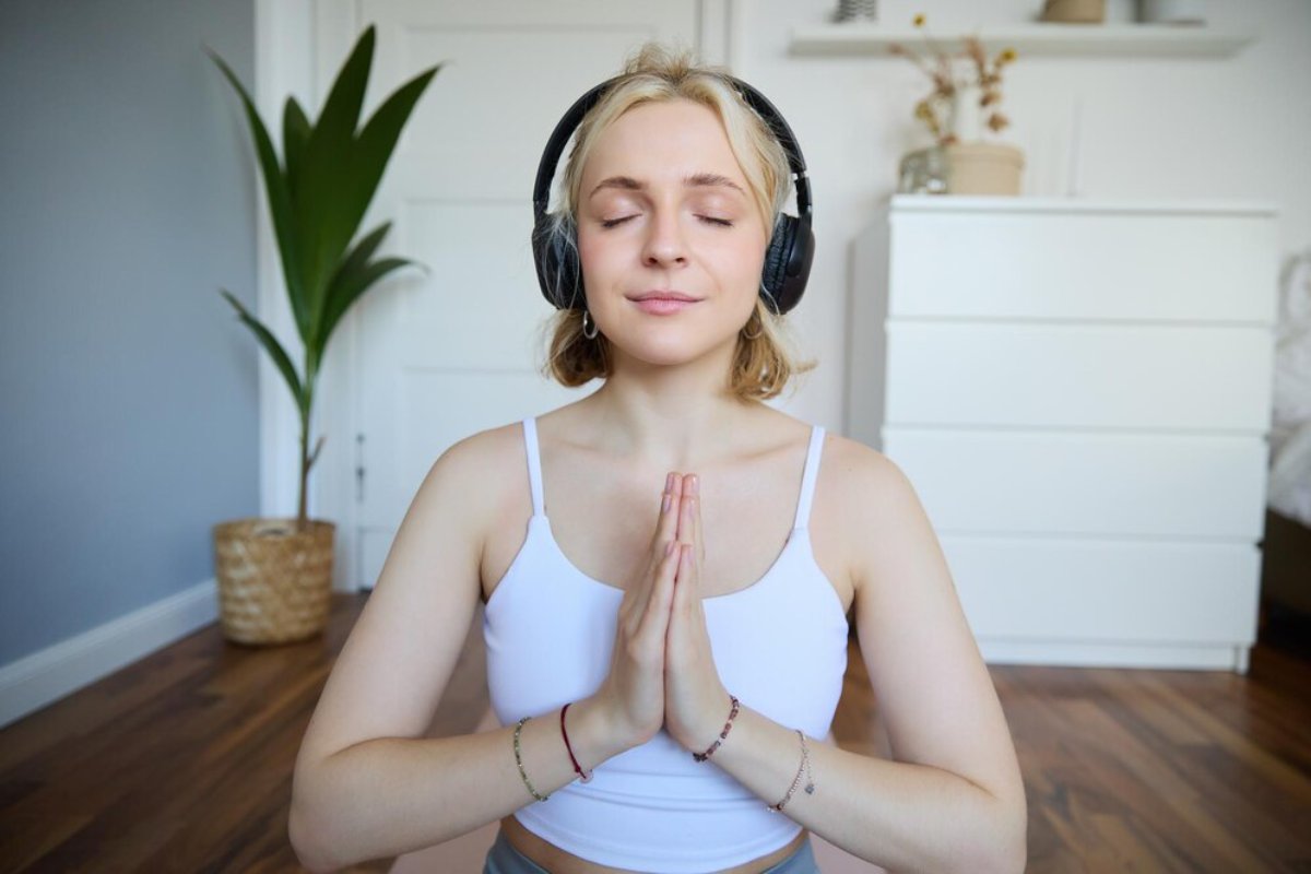 A person in a white tank top and headphones sits cross-legged, hands in a prayer position, surrounded by a serene indoor setting.