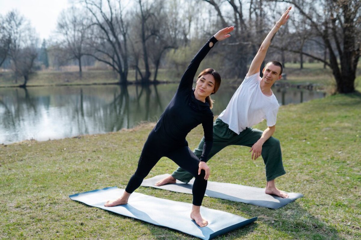 Two individuals practice yoga poses on mats by a serene lake, surrounded by trees in early spring.