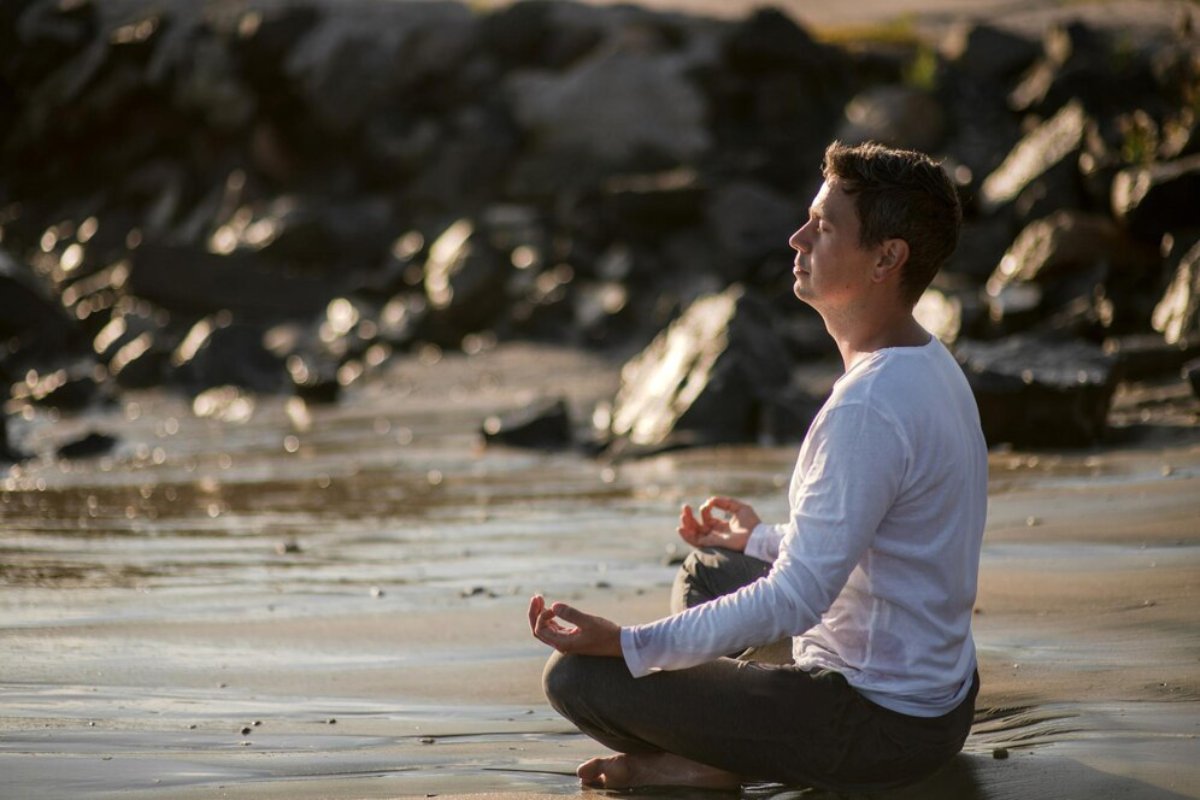 A person meditates cross-legged on a beach at sunset, surrounded by glistening water and dark rocks, creating a serene atmosphere.