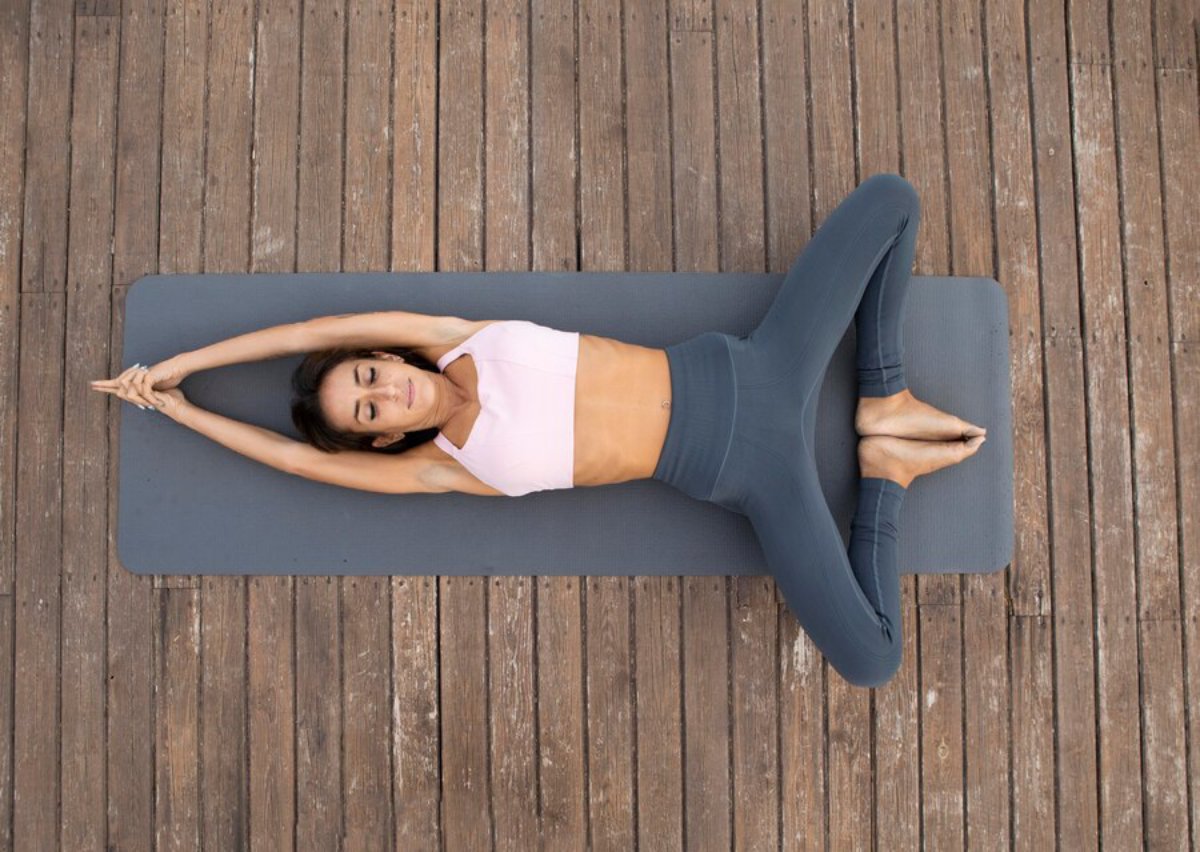 A person practicing yoga on a gray mat, stretching with arms extended overhead and legs spread wide on a wooden surface.