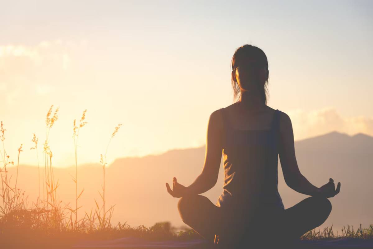 A silhouetted woman meditates outdoors at sunset, surrounded by grass and mountains, embodying tranquility and peace.
