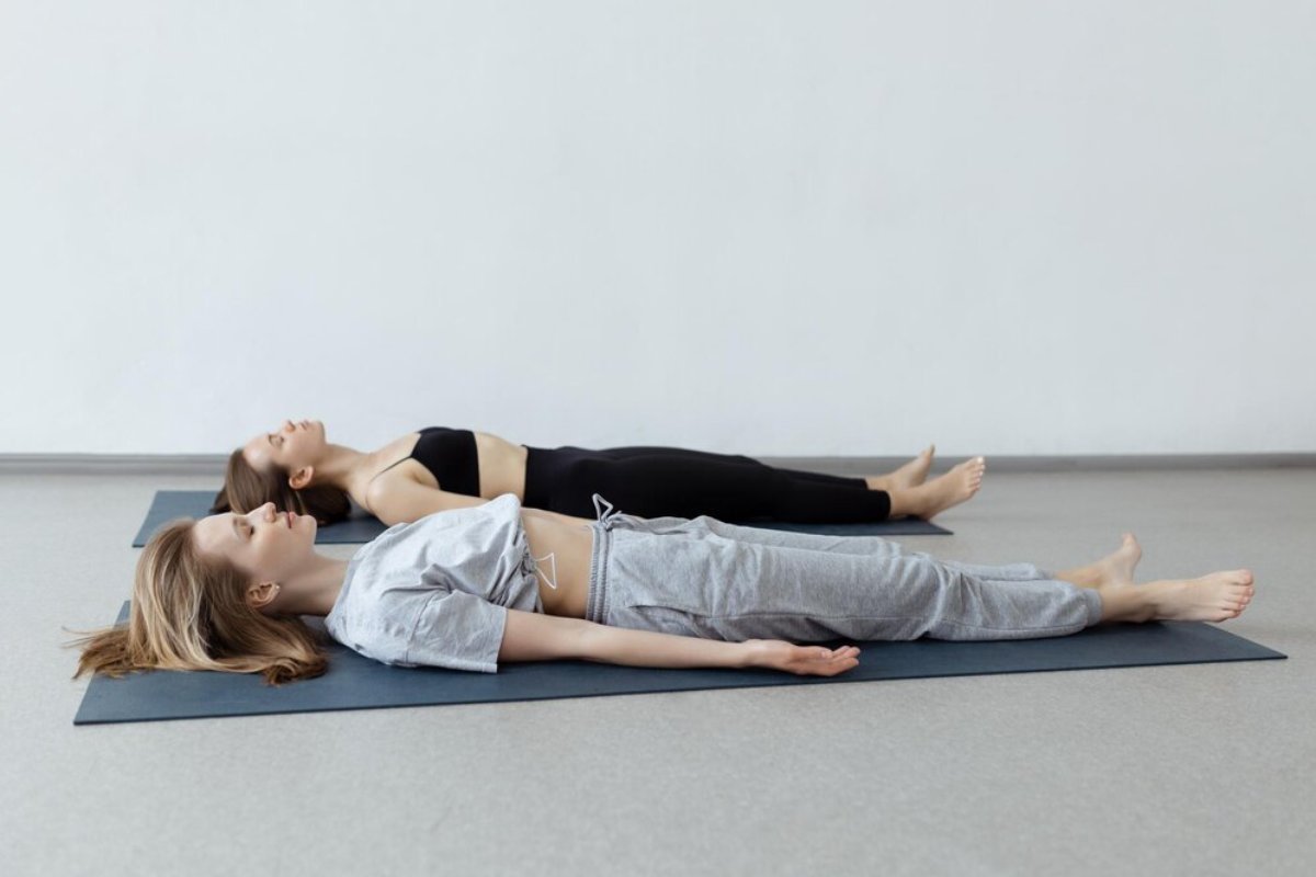 Two women practice yoga in a serene studio, lying on mats and relaxing in a restorative pose. Soft natural light fills the space.