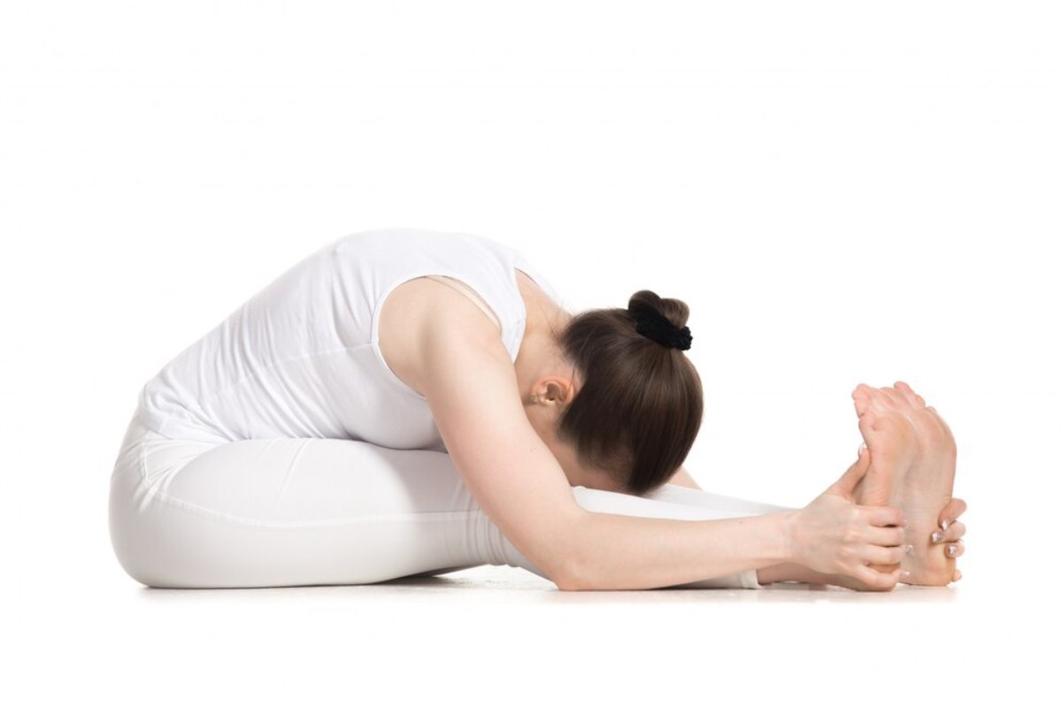 A woman in a white outfit stretches forward, grasping her feet while seated on the floor, demonstrating a yoga posture focused on flexibility.