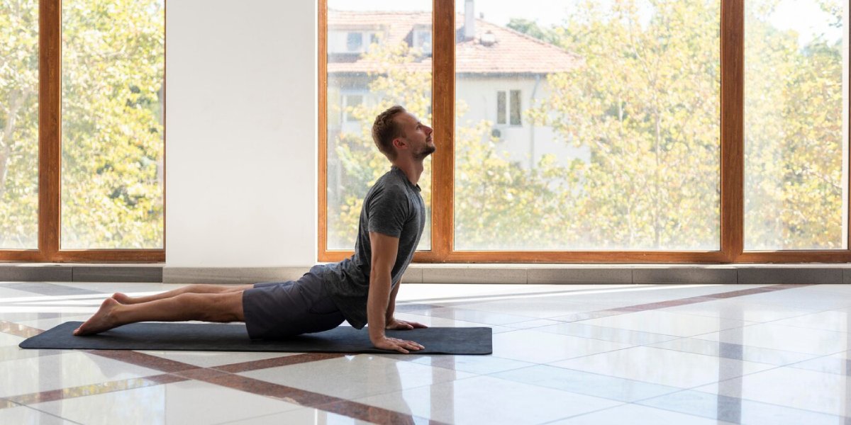 A person performs a yoga pose on a mat in a sunlit room with large windows and a view of greenery outside.