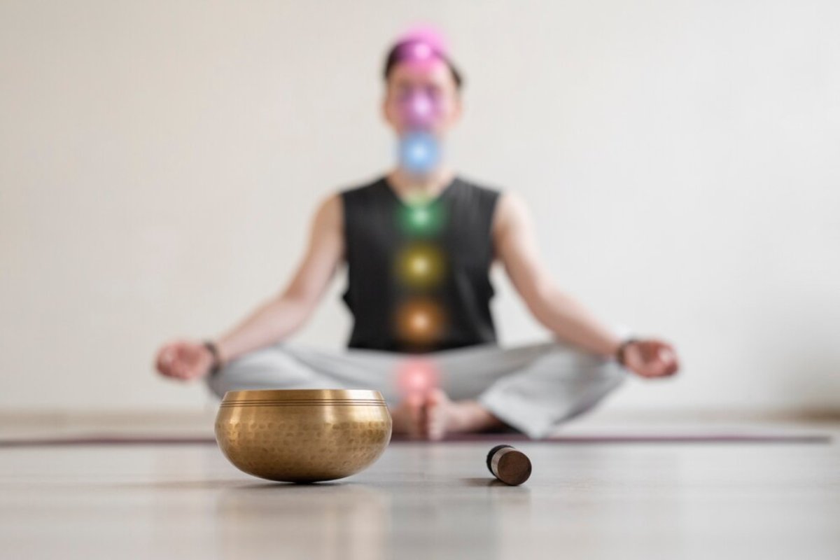 A person meditates with a singing bowl and mallet in the foreground, surrounded by colorful chakra lights in a serene space.