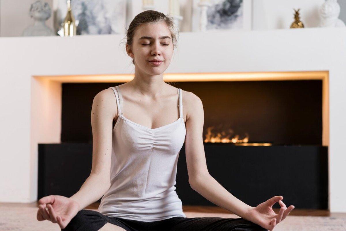 A person practices meditation in a modern living room, sitting cross-legged in front of a glowing fireplace.