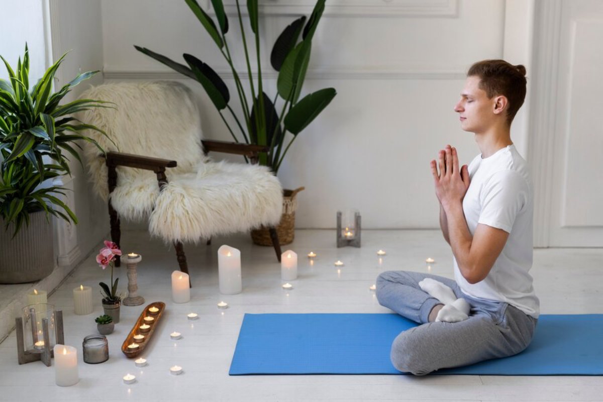 A person sits on a blue yoga mat, surrounded by candles and plants, creating a peaceful meditation space in a bright room.