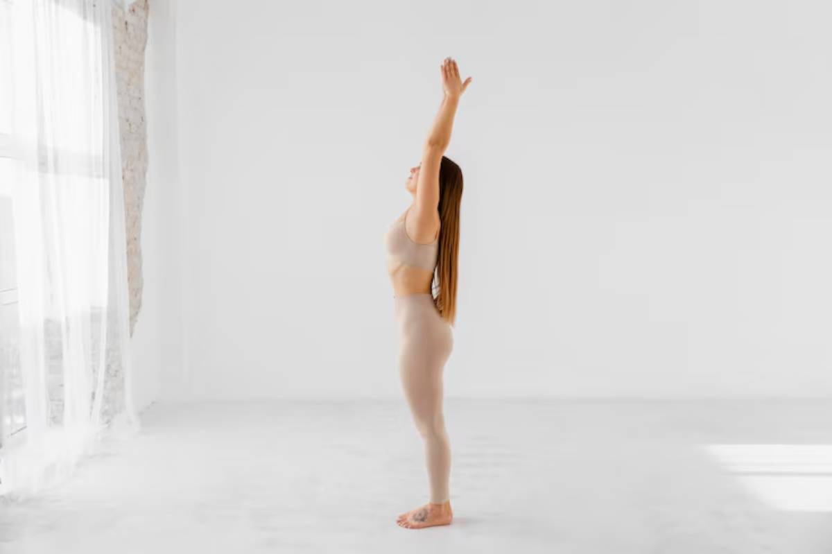 A woman in form-fitting beige workout attire stands upright with one arm raised, set against a bright, minimalistic indoor backdrop.