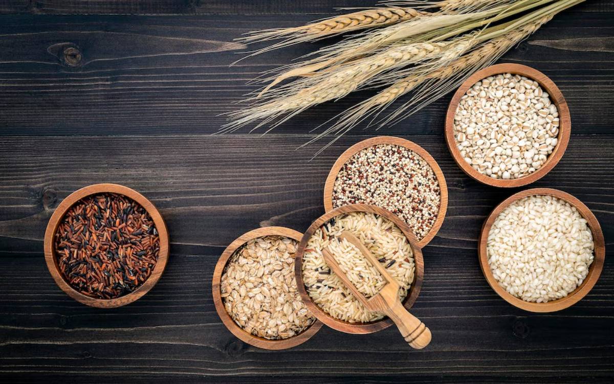 A rustic wooden surface displays bowls of various grains, including rice, oats, quinoa, and barley, alongside wheat stalks.