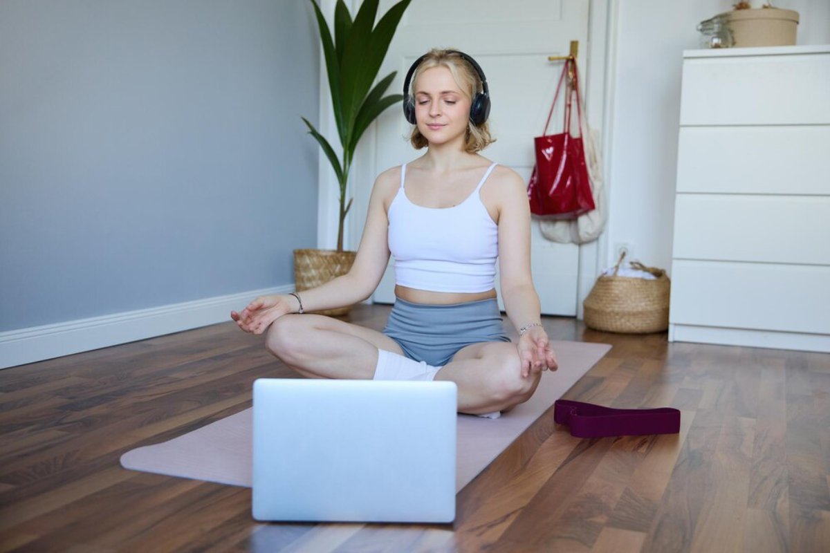 A woman in athletic wear meditates on a yoga mat in a serene room, with a laptop and plant nearby.