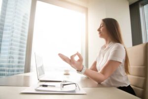 A person sits at a modern desk with a laptop, practicing mindfulness while looking out of a large window with city views.