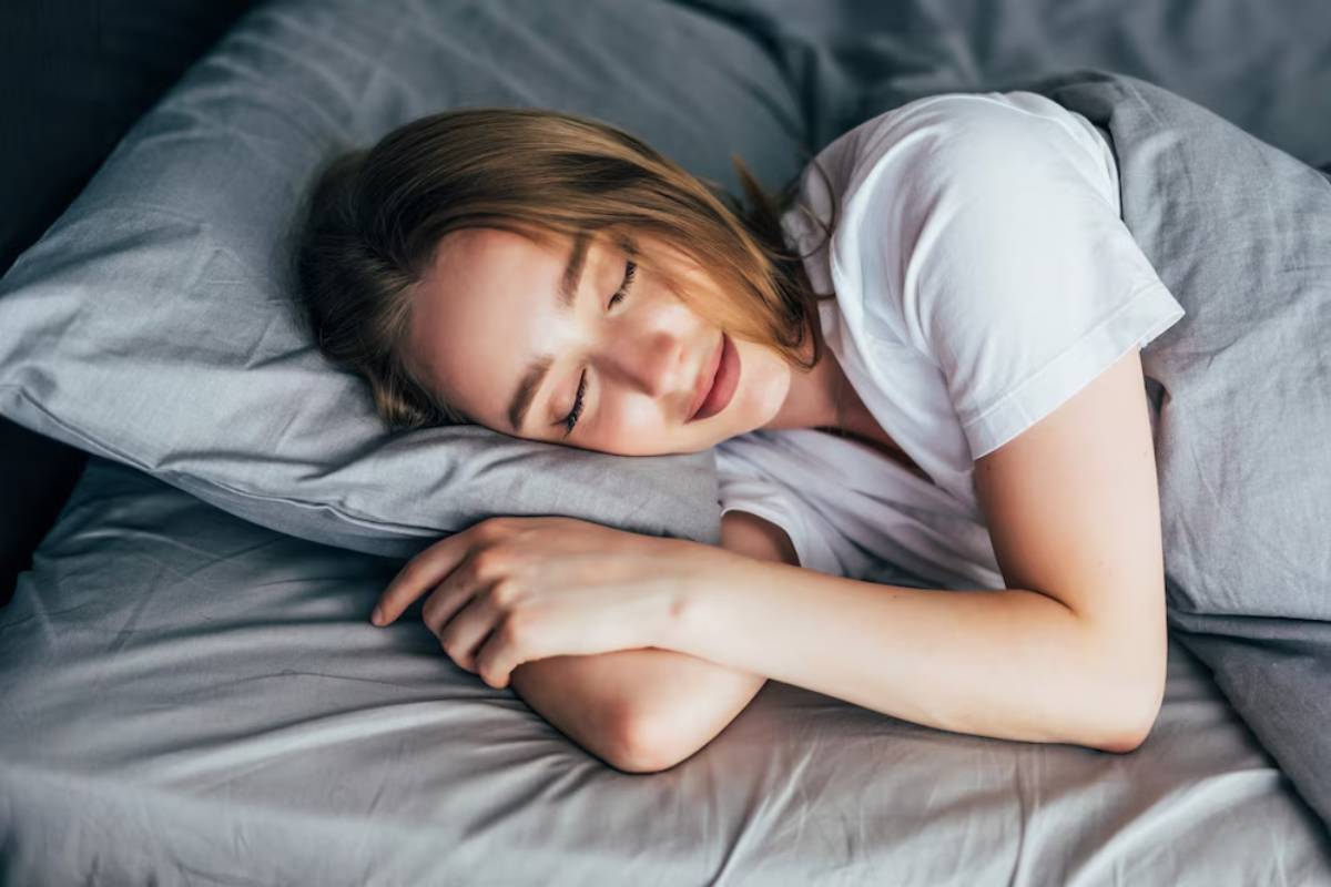 A person lies on a bed, resting their head on a pillow and curling their arms under their head, surrounded by soft gray bedding.