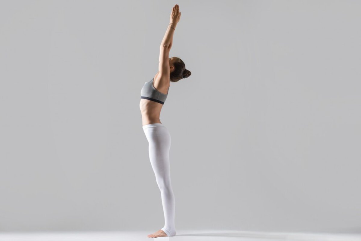 A woman in athletic wear performs a yoga pose, raising her arms overhead with a focused expression against a minimalist gray background.