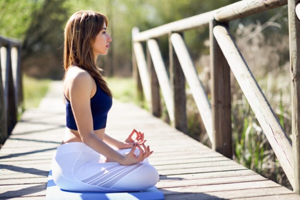 A person practices yoga on a blue mat, sitting cross-legged on a wooden pathway surrounded by greenery in bright sunlight.