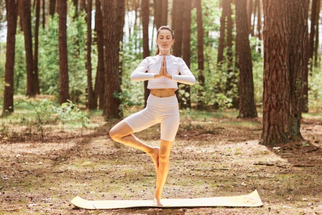 A person practicing yoga in a serene forest, balancing on one leg with hands in a prayer position, surrounded by tall trees.