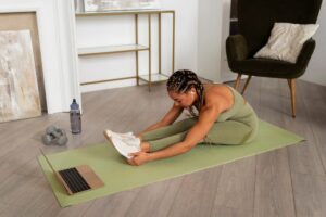 A person in activewear stretches on a green yoga mat at home, with a laptop, dumbbells, and a water bottle nearby.