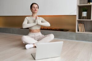 A woman sits cross-legged on a floor, practicing yoga in front of a laptop, with an elegant fireplace and bookshelf in the background.