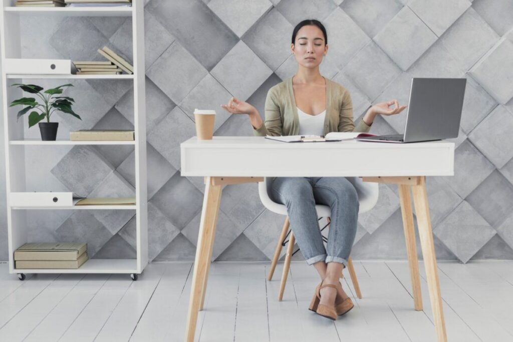 A person meditating at a modern desk with a laptop, coffee cup, and books, set against a stylish gray geometric wall.