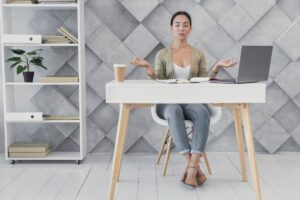 A person meditating at a modern desk with a laptop, coffee cup, and books, set against a stylish gray geometric wall.