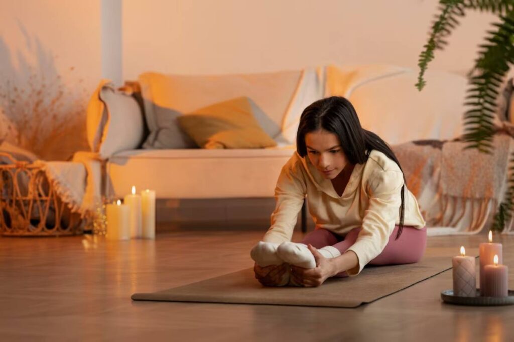 A person stretches on a yoga mat in a cozy living room with soft lighting, candles, and a couch with decorative pillows.