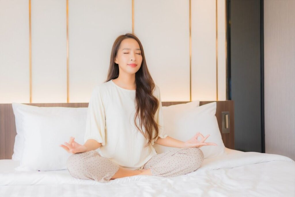 A woman sitting cross-legged on a bed, practicing meditation with a serene expression, surrounded by soft white pillows and warm lighting.