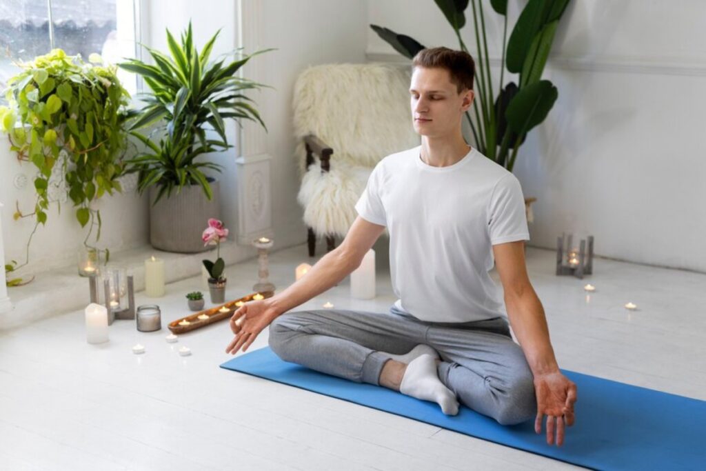 A person sits cross-legged on a yoga mat amidst plants and candles, practicing meditation in a serene indoor setting.
