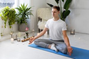 A person sits cross-legged on a yoga mat amidst plants and candles, practicing meditation in a serene indoor setting.